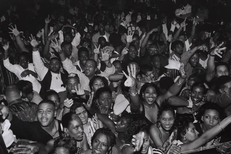 A lively black and white photograph of a crowded dance floor filled with people enjoying themselves, with hands raised and smiling faces celebrating.