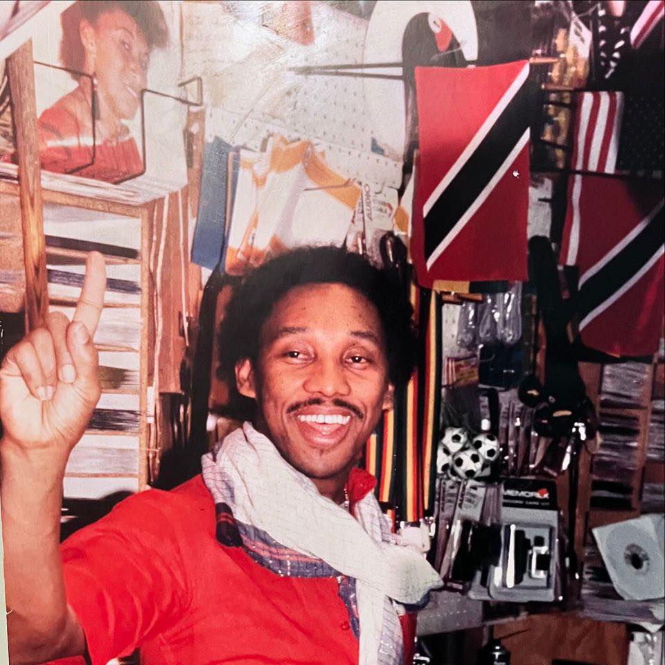 Smiling man wearing a red shirt and a scarf, raising his index finger, standing in a decorated store filled with various items and flags.