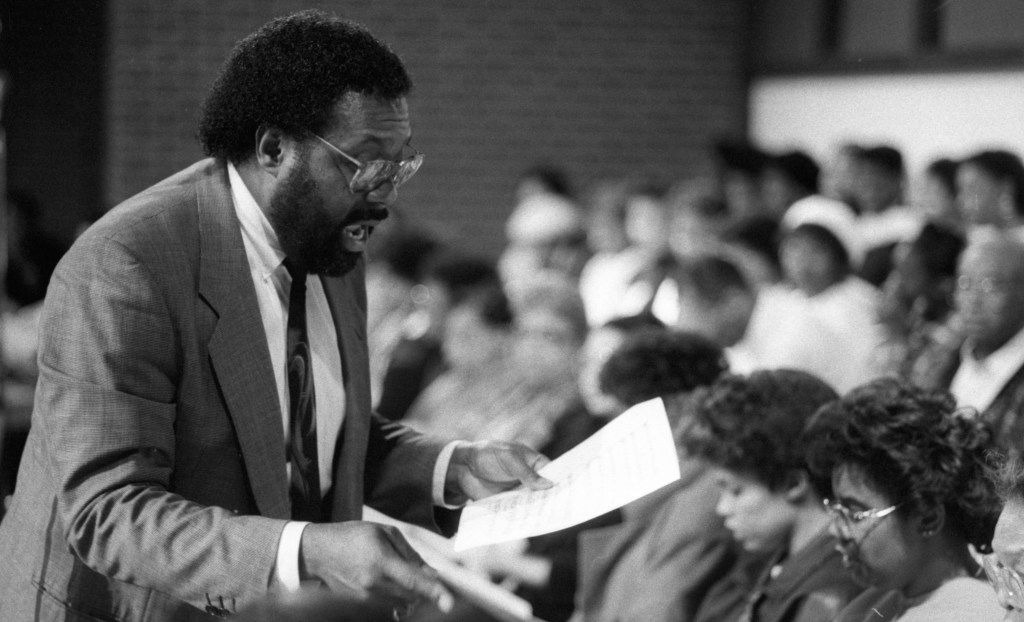 A speaker passionately addressing a crowd while holding a sheet of paper, surrounded by attentive audience members in a formal setting.
